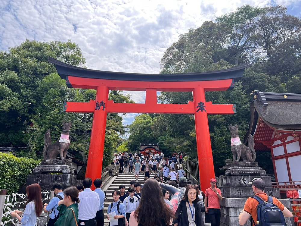 The main entrance of Fushimi Inari Taisha Shrine in Kyoto, dominated by a massive red torii gate and flanked by two stone fox (kitsune) statues wearing traditional bibs. A crowd of tourists is walking up the stone steps under a partly cloudy sky.
