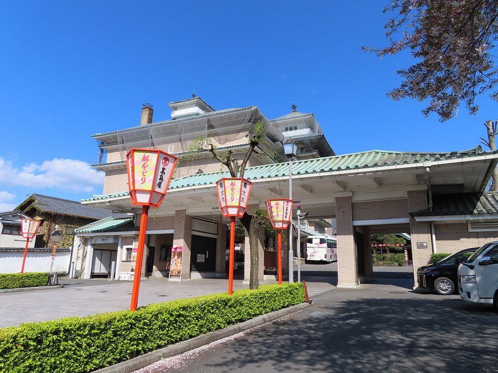 The entrance gate to the Gion Kobu Kaburenjo theater in Kyoto, featuring three tall, decorative red and white Japanese paper lanterns on stands. The lanterns feature floral motifs and Japanese characters for the Miyako Odori spring dances, set against a bright blue sky.

