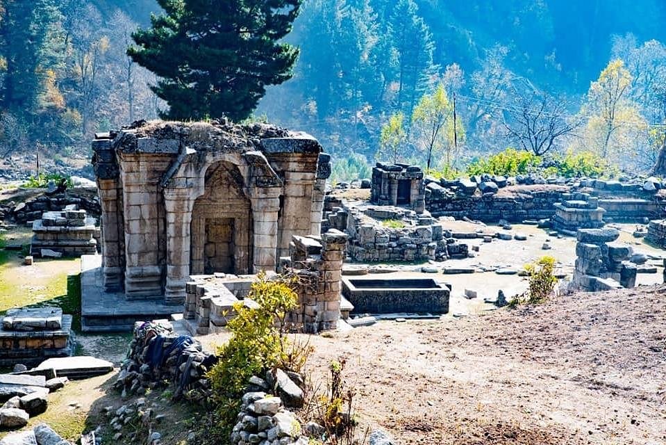 The historic Naranag Temple ruins in Ganderbal, Jammu and Kashmir. The photo captures the weathered grey stone architecture, including the main temple structure with its distinct Kashmiri gabled roof, carved stone walls, a rectangular stone water tank (kund), and a background of dense pine trees on a sunny hillside.