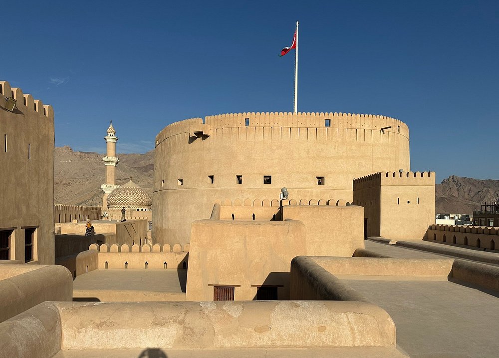 The majestic, sand-colored round tower of Nizwa Fort in Oman, featuring a red flag flying on a tall flagpole, with courtyard rooftops and a mosque dome and minaret visible in the background under a clear blue sky.