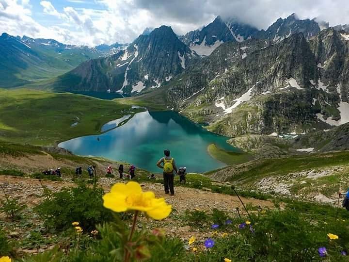 Scenic view from the Kashmir Great Lakes Trek showing the turquoise waters of Kishansar Lake cradled by steep, rocky mountain peaks with lingering snow. A hiker wearing a lime-green backpack stands on the trail overlooking the water, with wild yellow flowers blooming in the grassy foreground under a cloudy sky.