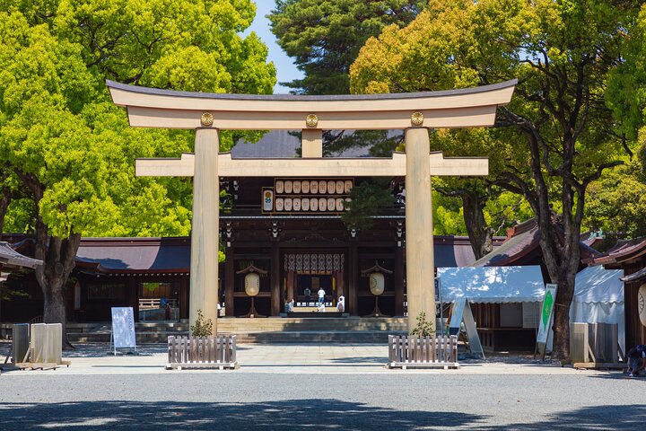 A grand, light-colored wooden torii gate standing at the entrance to a traditional Japanese Shinto shrine, surrounded by mature, leafy green trees.