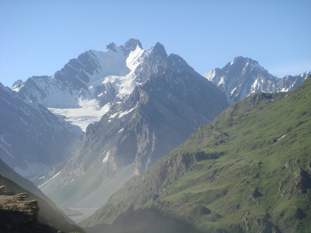 A majestic view of the sharp, snow-covered Machoi Peak and its active glaciers under a clear blue sky. A vibrant green, grassy mountain slope cuts diagonally across the foreground, contrasting with the icy, rugged terrain of the Himalayan range near Sonamarg and Zoji La.