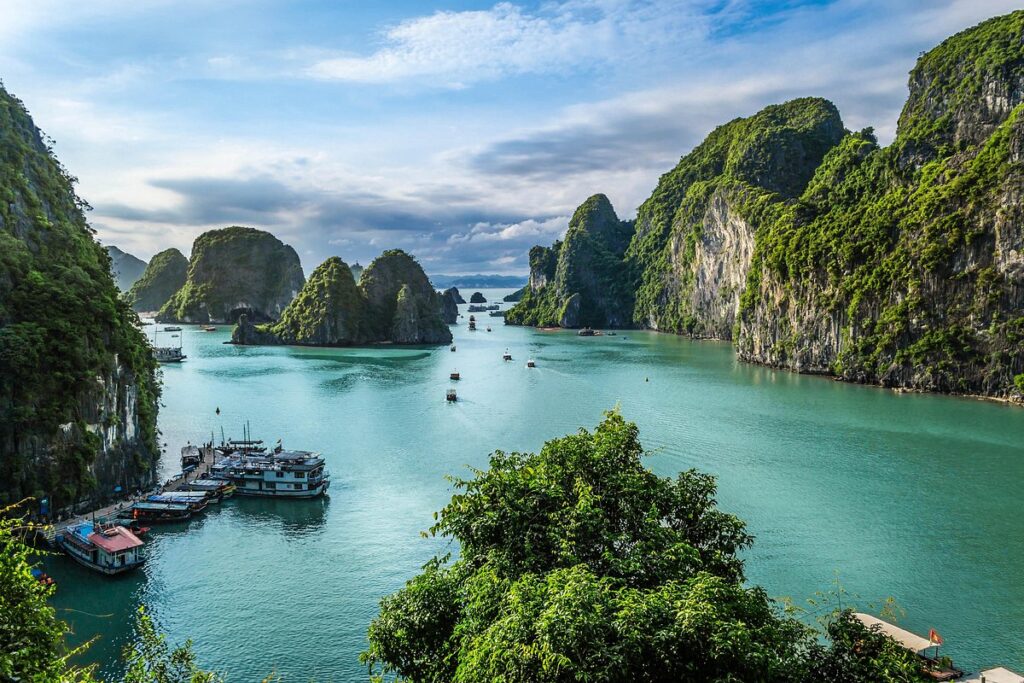 Scenic view of Ha Long Bay in Vietnam, featuring towering emerald-green limestone karsts rising from calm, turquoise waters with several tourist cruise boats.