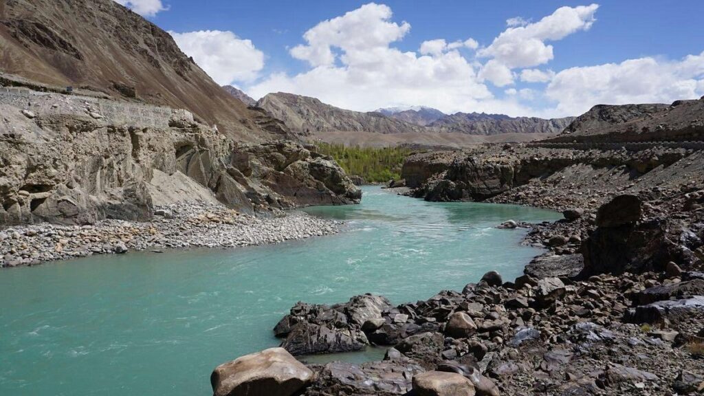 Indian Himalayas. The turquoise waters of a glacial river winding through a steep valley surrounded by pine-forested slopes and distant peaks.