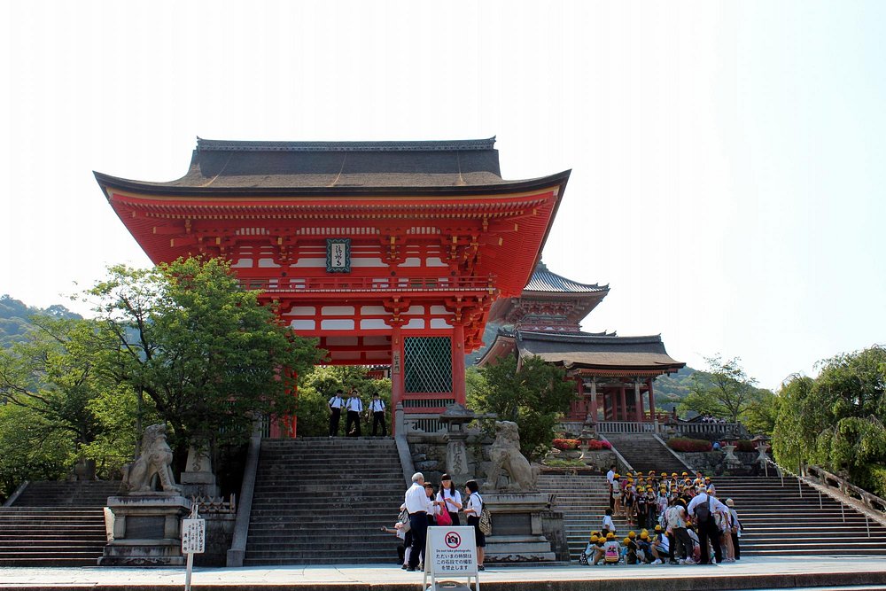 The striking red and orange Niomon gate of Kiyomizu-dera Temple in Kyoto, Japan. Visitors and school children are gathered on the wide stone staircase leading up to the grand entrance, which is guarded by stone komainu (lion-dog) statues.
