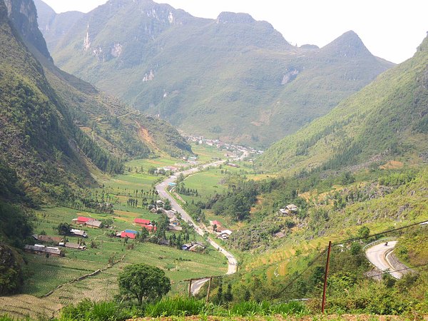 Lush green mountain valley in Northern Vietnam with a winding paved road leading past rural villages and agricultural fields surrounded by steep hills.