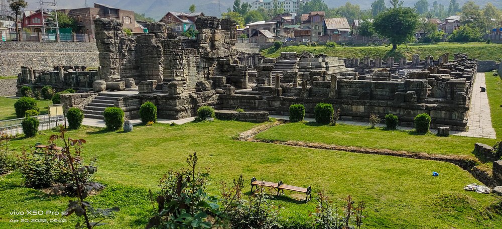 The ancient grey stone ruins of the 9th-century Avantiswami Temple in Awantipora, Kashmir, situated in a green manicured lawn with small rose bushes and residential houses in the background.