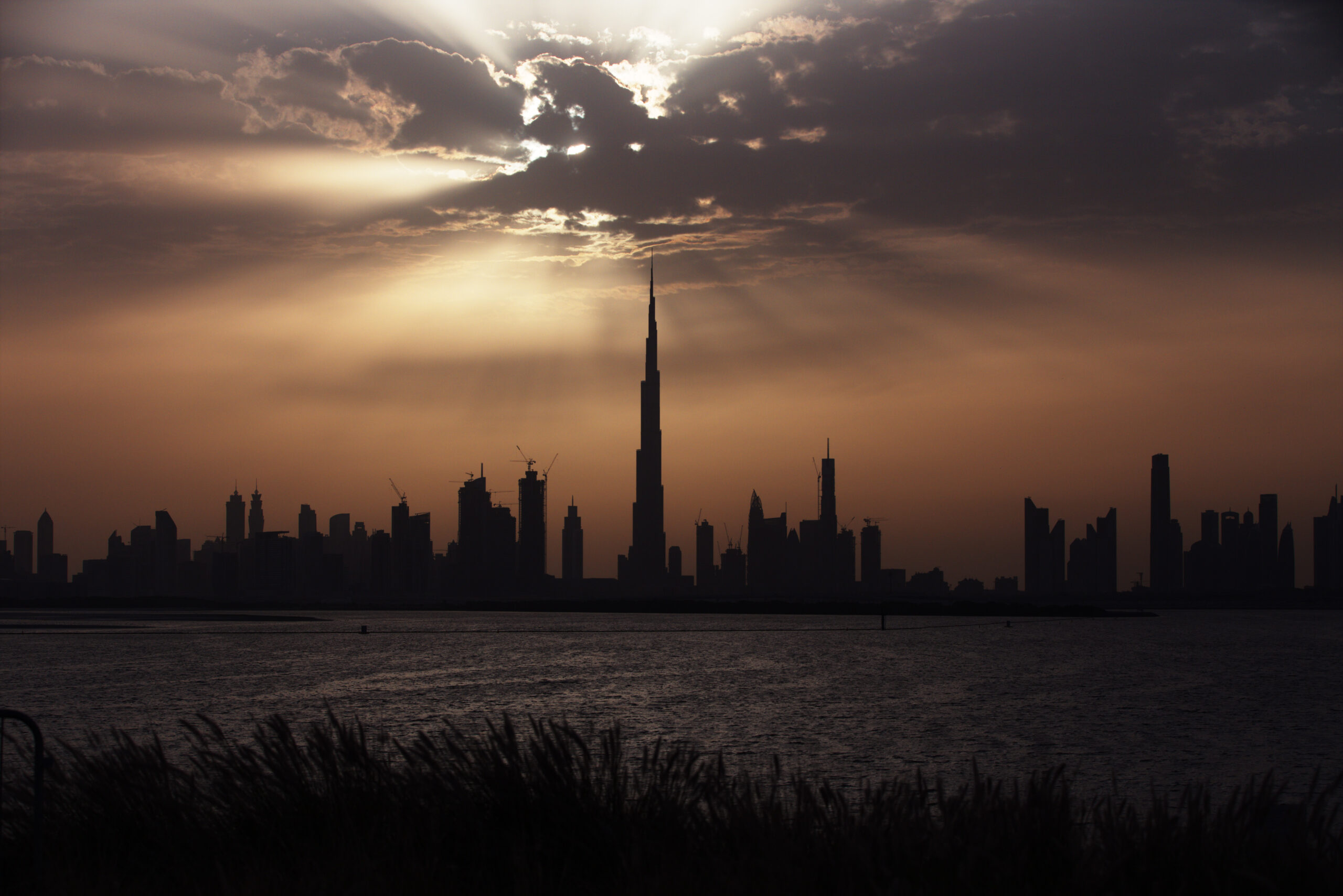 The towering Burj Khalifa skyscraper reaching into a blue sky with soft white clouds, surrounded by the modern Downtown Dubai skyline, green lawns, and local streets.