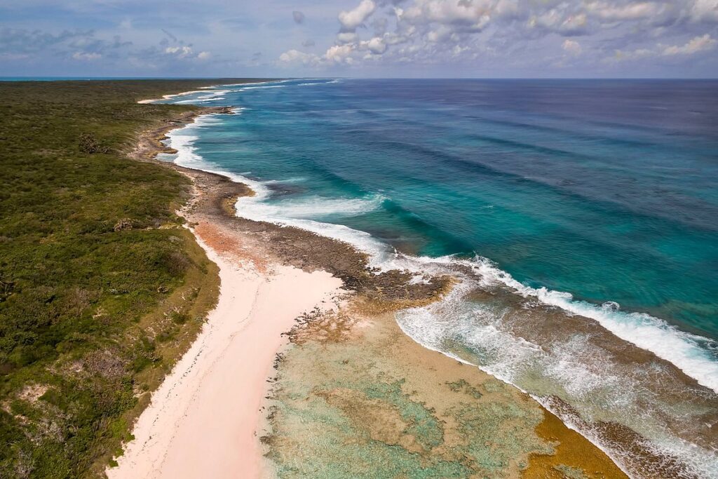 An aerial, high-angle view of a tropical coastline showing vibrant turquoise ocean waves breaking over a shallow coral reef. A wide, light-colored sandy beach stretches along the shore, bordered on the left by a dense, lush green forest under a partly cloudy blue sky.