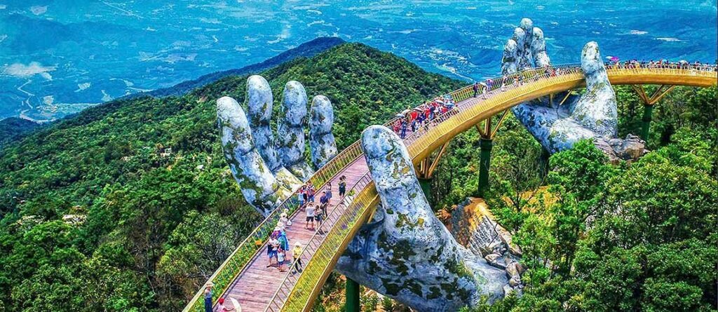 An aerial, wide-angle view of the Golden Bridge (Cầu Vàng) in the Ba Na Hills near Da Nang, Vietnam. Tourists stroll along the curved, golden pedestrian bridge which is held aloft by two giant, weathered, moss-covered stone hands. The bridge is surrounded by dense, lush green mountain forests stretching into the distant valley below.