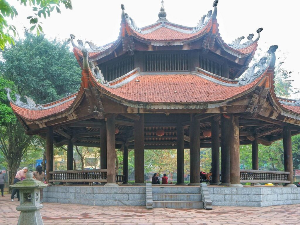 A close-up shot of the traditional Vietnamese octagonal wooden pavilion located in Giam Garden at the Temple of Literature in Hanoi. The open-air wooden structure features a double-tiered, red clay-tiled roof with elegantly curved eaves, supported by thick, round dark wooden pillars resting on a low stone platform.