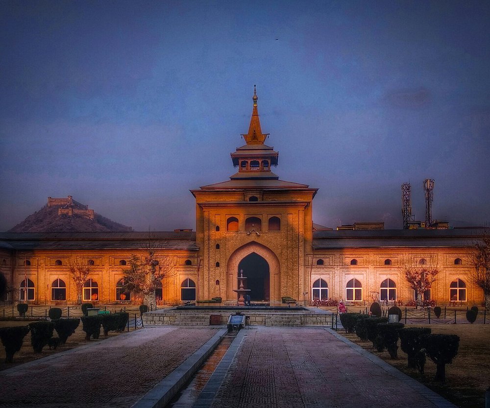 Symmetrical view of a historic brick building with traditional Kashmiri architecture and a multi-tiered pagoda spire, illuminated by warm orange lights at dusk, with Hari Parbat Fort visible on a distant hill under a deep blue sky.