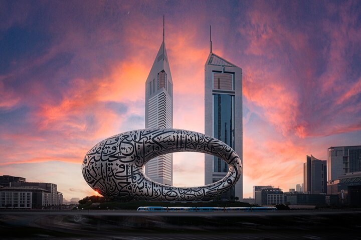 The towering Burj Khalifa skyscraper reaching into a blue sky with soft white clouds, surrounded by the modern Downtown Dubai skyline, green lawns, and local streets.
