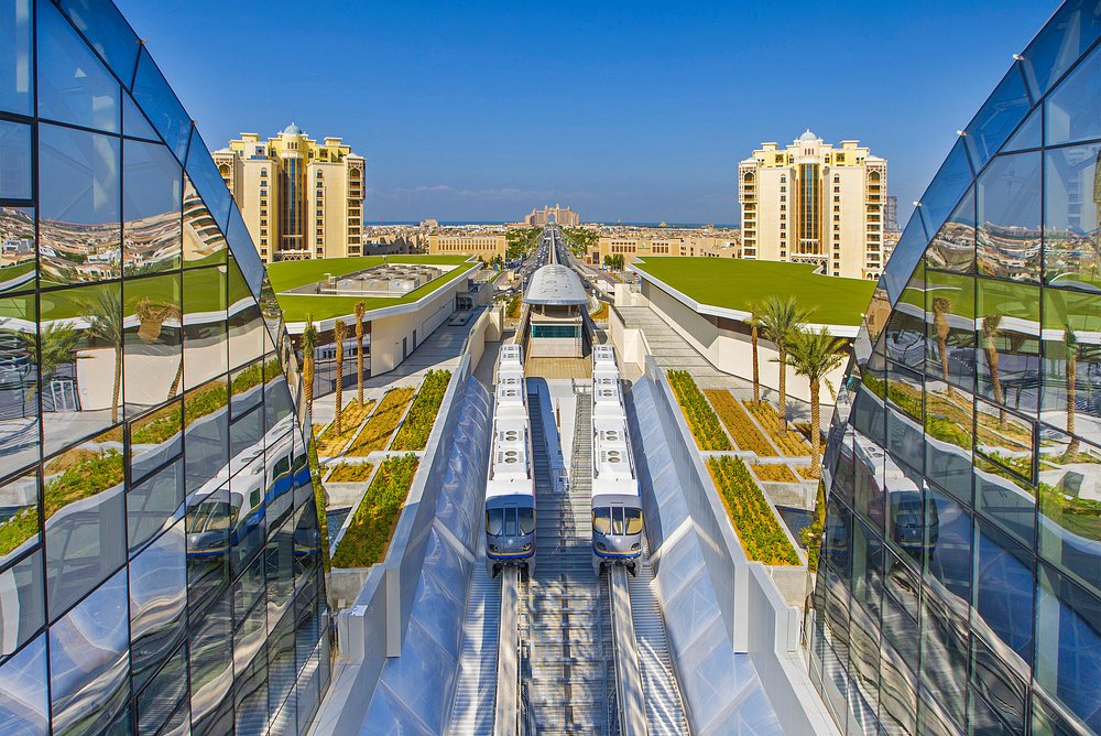 A symmetrical, high-angle view of the Palm Jumeirah Monorail tracks in Dubai, framed by modern glass-faced buildings with the iconic Atlantis, The Palm hotel visible in the far distance.

