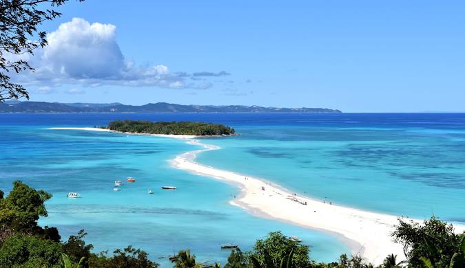 Turquoise waters lapping onto a pristine white sand beach in [Location, e.g., Nosy Be, Madagascar], with palm trees swaying in the breeze.