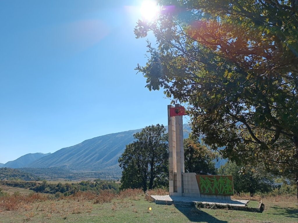 A stone monument featuring the Albanian flag and green figures stands in a grassy field in Zagoria Valley. A large tree frames the monument, and mountains are seen under a bright sunny sky.