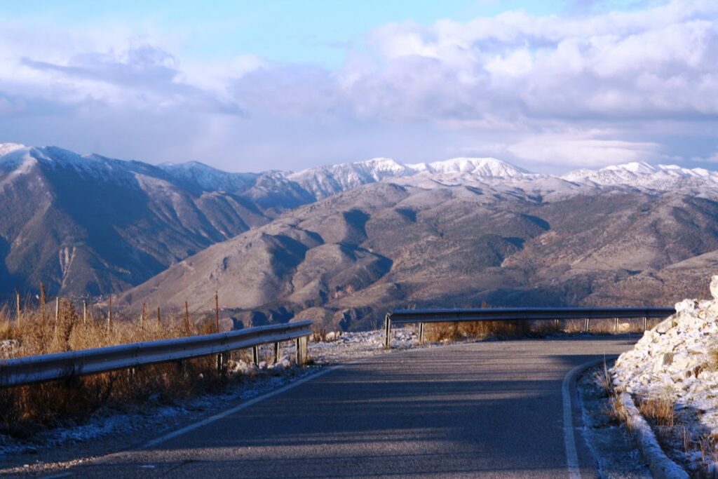 A paved road curves through a mountainous region of Zagoria Valley. Snow-capped peaks are visible in the distance under a partly cloudy sky, with patches of snow along the roadside.