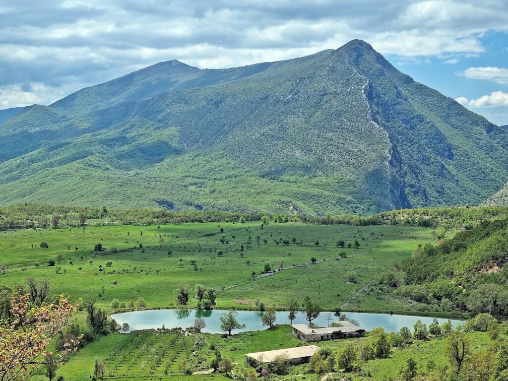 A panoramic view of a lush green valley in Zagoria, Albania, featuring a small lake reflecting the surrounding landscape. A prominent mountain peak dominates the horizon under a partly cloudy sky.
