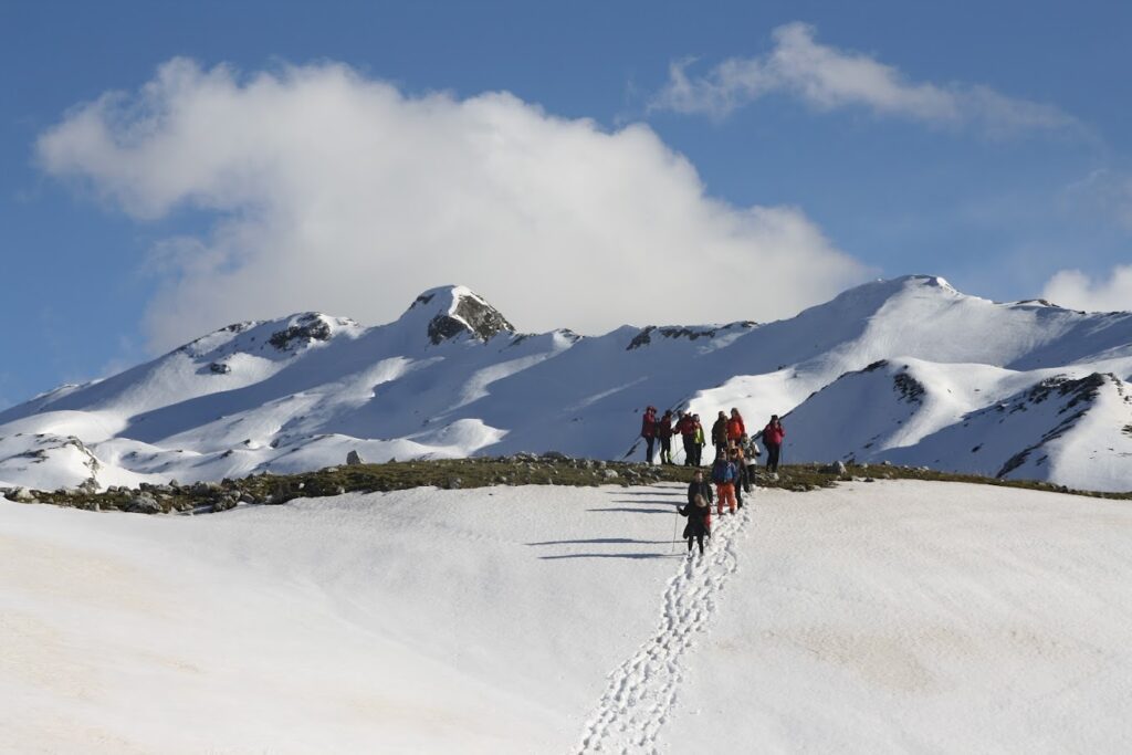 A group of hikers trekking through a snowy landscape in Zagoria Valley, Albania, with mountains rising in the background under a blue sky with white clouds. Footprints are visible in the snow leading up a gentle slope.
