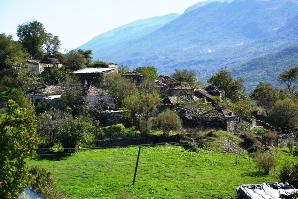 A traditional Albanian village with stone houses and tiled roofs nestled on a green hillside in Zagoria Valley. Lush vegetation surrounds the village, and mountains are visible in the background.