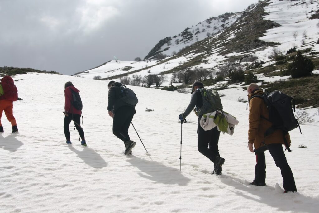 A group of hikers trekking through a snowy landscape in Zagoria Valley, Albania, with mountains rising in the background under a blue sky with white clouds. Footprints are visible in the snow leading up a gentle slope.