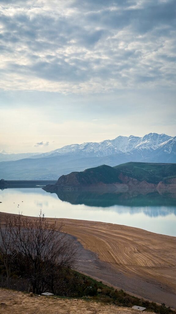 Tranquil lake reflecting snow-covered mountains under a cloudy sky in Uzbekistan.