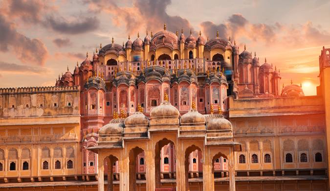 High-angle view of the City Palace in Jaipur, India. The image highlights the palace's stunning architecture, featuring a blend of red and yellow sandstone structures with multiple domes and detailed facades. People can be seen on the upper terraces of the palace.