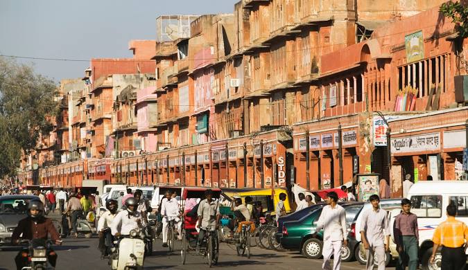 Johari Bazaar. A vibrant street scene in Jaipur's old city, with people walking and riding various modes of transportation, passing by traditional terracotta-colored buildings filled with shops and activity. 