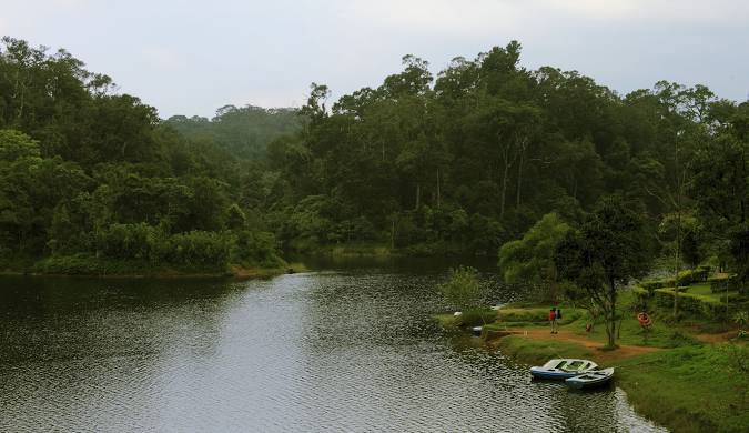 Serene Gavi Lake in Kerala, surrounded by lush green forests, with two boats resting on the shore.