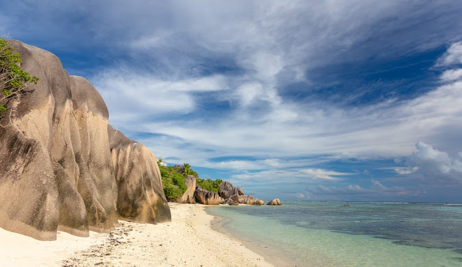 Wide panoramic view of a Seychelles beach featuring distinctive granite boulders, white sand, and the turquoise Indian Ocean stretching to the horizon under a partly cloudy sky. This is the kind of scenery featured in magazine articles about travel, especially to Seychelles.