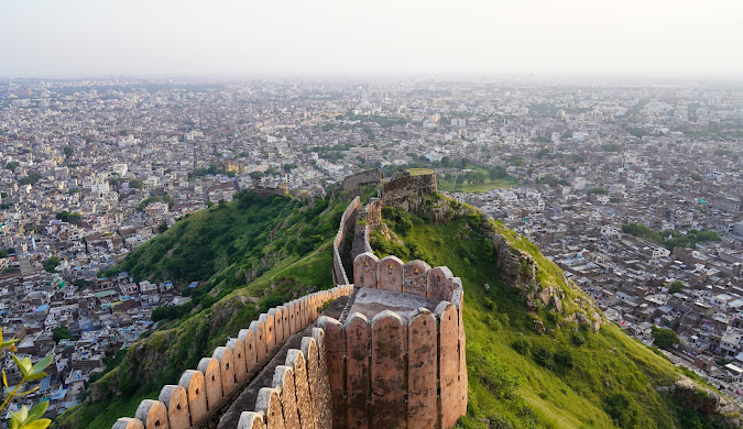 Aerial view of a weathered stone fort wall winding along a hillside, overlooking a sprawling city. The wall is terracotta-colored with battlements.
