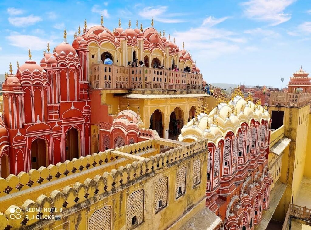 High-angle view of the City Palace in Jaipur, India. The image highlights the palace's stunning architecture, featuring a blend of red and yellow sandstone structures with multiple domes and detailed facades. People can be seen on the upper terraces of the palace.