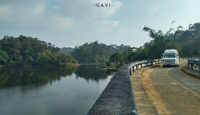 A winding road along the edge of Gavi Lake in Kerala, with a white tourist van navigating the curve, offering picturesque views of the tranquil waters and surrounding forest.
