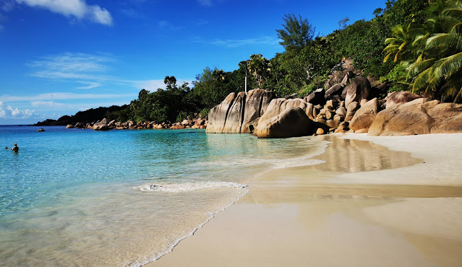 Wide panoramic view of a Seychelles beach featuring distinctive granite boulders, white sand, and the turquoise Indian Ocean stretching to the horizon under a partly cloudy sky. This is the kind of scenery featured in magazine articles about travel, especially to Seychelles.