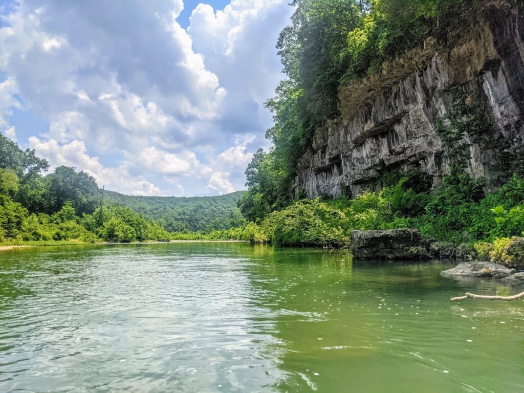 Kayakers paddling on a clear, green river beneath limestone bluffs in Mark Twain National Forest. Experience the dramatic scenery of the Ozarks while Missouri River rafting Ozarks, with towering bluffs and lush vegetation.